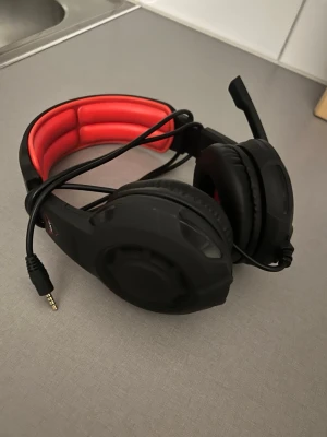 Svarta gaminghörlurar med mikrofon - Säljer ett par svarta gaminghörlurar med röd vadderad bygel och inbyggd mikrofon. Hörlurarna har over-ear design och ansluts med 3,5 mm kabel. Perfekta för gaming eller videosamtal, med bekväm passform och robust konstruktion.