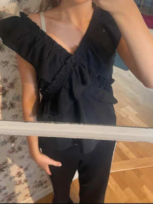 Svart omlottopp med volanger - Snygg svart omlottopp med breda volanger och djup v-ringning. Toppen är ärmlös och har en luftig passform som ger en cool och trendig look. Perfekt att styla med jeans eller kjol för en chic vibe.