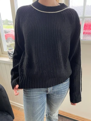 Svart stickad tröja med vit detalj - Säljer en svart ribbstickad tröja med vit kontrastlinje runt halsen och längs ärmarna. Tröjan har en avslappnad passform, rund halsringning och långa ärmar.