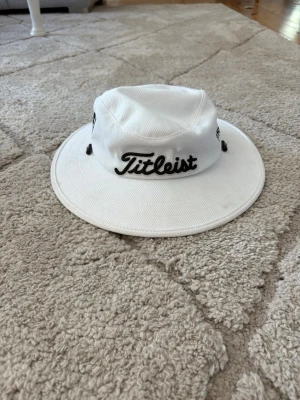 Vit solhatt från Titleist - Snygg vit solhatt från Titleist med bred skärm och svart logga broderad fram och bak. Hatten har justerbar dragsko för perfekt passform och är tillverkad i ett lätt och ventilerande material. Perfekt för soliga dagar på golfbanan eller stranden.