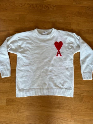 Vit stickad tröja från ami Paris - Vit stickad tröja med rund hals och ribbade muddar. På bröstet finns ami Paris loggan, vilket ger en unik och trendig look. Perfekt för dig som gillar stilrena plagg med en twist.