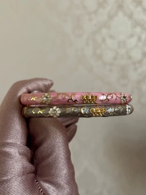 Enamel bangles - Två armband, ett i rosa och ett i grått. Armbanden har en rund form och är tillverkade i ett hårt material med emaljliknande yta. 