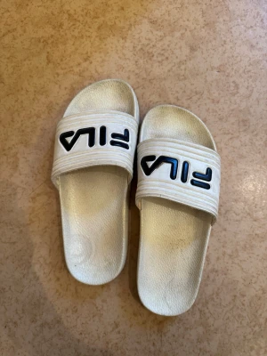 Vita Fila tofflor - Vita tofflor från Fila med bred rem och svart logga framtill. Klassisk slip-in modell i syntetmaterial, perfekt för chill dagar hemma eller utomhus. 