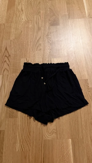 Svarta shorts med snörning - Säljer ett par svarta shorts med hög midja och snörning i midjan. Shortsen har en avslappnad passform och är tillverkade i ett mjukt, tunt tyg som passar perfekt till varmare dagar. Midjan är elastisk och har små dekorativa hjärtan på snöret. Köpta på newyorker, står att det är storlek M men dom är mer Xs/Xxs. Om inte ännu mindre nästan, så dom är väldigt små.                                             Katt finns i hemmet!!