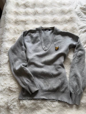 Grå V-ringad tröja Lyle & Scott - Snygg grå V-ringad tröja från Lyle & Scott med den klassiska gula loggan på bröstet. Tröjan har långa ärmar, ribbade muddar och är tillverkad i mjukt material som passar perfekt till lager på lager. Enkel och stilren design. Den har några skavanker, men inget som inte går att fixa. Säljer även på Vinted @caisaborelius