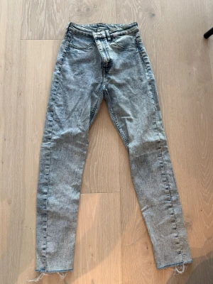 Grå tvättade skinny jeans - Säljer ett par grå skinny jeans med tvättad look och råa, fransiga benslut. Jeansen har klassisk femficksdesign och hög midja. Perfekta för en avslappnad och trendig stil.