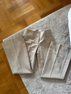 Beige kostymbyxa med hög midja - Snygga beige kostymbyxor med hög midja och raka ben. Byxorna har pressveck framtill, bred linning och dubbla knappar. Materialet ser ut att vara en mjuk och lite stretchig blandning, perfekt för en clean och stilren look. Storlek s/xs