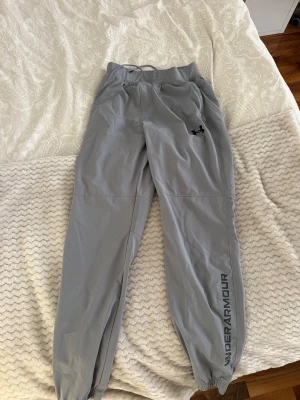 Grå mjukisbyxor från Under Armour - Snygga grå mjukisbyxor från Under Armour med resår i midjan och dragsko. Byxorna har logga på låret och texten UNDER ARMOUR längs benet. Tillverkade i ett lätt och mjukt material, perfekta för chill eller träning.