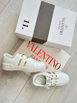 Vita Valentino sneakers med nitar - Snygga vita sneakers från Valentino med guldfärgade nitar på sidorna och över vristen. Skorna har en klassisk låg siluett, vita snören och är tillverkade i slätt läder. Perfekta för dig som vill sticka ut med exklusiva detaljer.