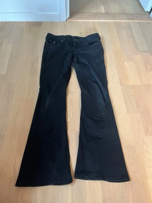 Svarta True Religion flare jeans - Säljer ett par svarta flare jeans från True Religion, modell Joey Low Rise Flare. Jeansen har låg midja, utsvängda ben och klassisk femficksdesign. De köptes i vintras så de är relativt nya. Skrevet har lite slitningar men det är inget som syns. De är i W27