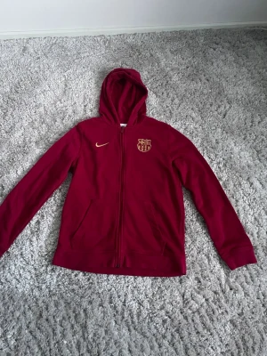 Vinröd FC Barcelona hoodie med dragkedja - Snygg vinröd hoodie från Nike med FC Barcelona-logga på bröstet och gul Nike-swoosh. Hoodien har dragkedja framtill, två fickor och en klassisk huva. Perfekt för dig som älskar fotboll och vill ha en sportig look.