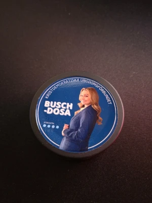 Ebba Busch-dosa - En svart snusdosa med blått lock och tryck av en kvinna i blå kavaj samt texten 'BUSCH-DOSA'. På locket står även 'Kristdemokratiska Ungdomsförbundet'. Dosan är rund och gjord av plast.