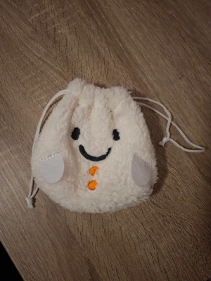 Söt fluffig påsväska med ansikte - En liten, mjuk och fluffig påsväska i vitt med dragsko. Väskan har ett broderat ansikte med svarta ögon och leende, samt orange detaljer och vita applikationer på sidorna. Perfekt för småprylar eller accessoarer.