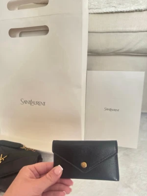 Svart plånbok från Saint Laurent - Snygg svart plånbok från Saint Laurent i slätt läder med guldfärgad knappstängning. Plånboken har en stilren och minimalistisk design i kuvertmodell, perfekt för kort och sedlar. En lyxig accessoar som lyfter din stil.