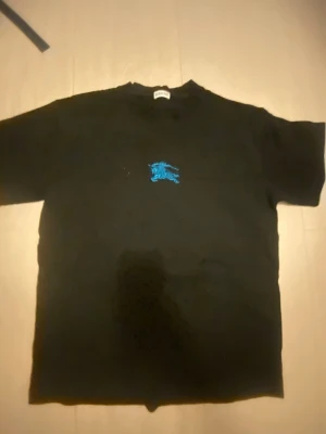 Svart Burberry t-shirt med blå logga - Säljer en svart t-shirt från Burberry med en blå broderad logga på bröstet. Klassisk rund halsringning och korta ärmar. Tillverkad i mjuk bomull för skön känsla. Perfekt för dig som gillar stilrena plagg med en twist.