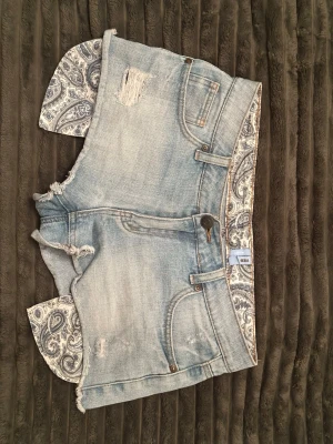 Ljusblå jeansshorts med paisleydetaljer - Säljer ett par ljusblå jeansshorts med slitna detaljer och paisleymönstrade tygkanter som sticker ut nedtill. Shortsen har fem fickor, knappgylf och en avslappnad passform. Perfekta för sommaren och ger en cool, unik look.