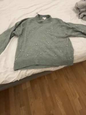 Grå stickad tröja med pärlor H&M - Mysig grå stickad tröja från H&M i storlek XS. Tröjan har rund hals, ribbade muddar och är dekorerad med små pärlor över hela framsidan för en extra snygg touch. Perfekt för dig som gillar en enkel men ändå trendig look.