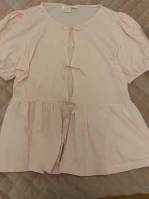 Ljusrosa blus med knyt och puffärm - Söt ljusrosa blus med korta puffärmar och tre dekorativa knyt framtill. Blusen har en lös passform och lätt volang nedtill, vilket ger en romantisk och trendig look. Perfekt att styla med jeans eller kjol för en feminin vibe.