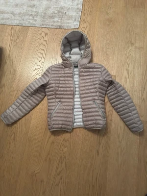 Beige lättviktsdunjacka med huva - Snygg beige dunjacka med quiltad design och fast huva. Jackan har två dragkedjeförsedda fickor framtill och diskret logga på ärmen. Perfekt för kalla vinterdagar och enkel att matcha med olika outfits. Passar S-M 