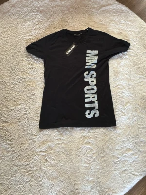Svart t-shirt med tryck från MM Sports - Snygg svart t-shirt från MM Sports med stort vitt tryck både fram och bak. Klassisk passform och rund halsringning. Perfekt för dig som gillar sportig stil och vill ha en enkel men cool t-shirt i garderoben.