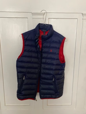 Mörkblå dunväst från Polo Ralph Lauren Junior L 14/16 - Snygg mörkblå dunväst från Polo Ralph Lauren med röd insida. Västen har två dragkedjeförsedda fickor och klassisk broderad logga på bröstet. 