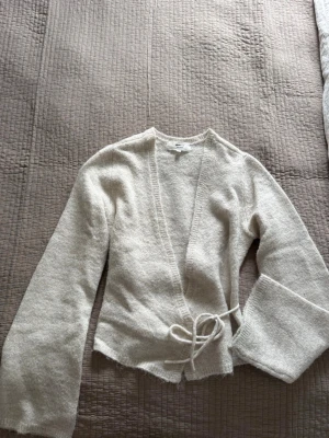Beige omlottkofta från Gina Tricot - Mysig beige stickad omlottkofta från Gina Tricot i ullblandning. Koftan har långa, lite vidare ärmar och knyts i midjan med ett band. Perfekt för lager på lager och ger en avslappnad men ändå stilren look.