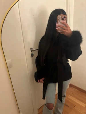 Svart kappa med fuskpälsdetaljer - Snygg svart kappa med bälte i midjan och fluffiga fuskpälsdetaljer vid ärmsluten. Rak passform  som ger en trendig siluett. Perfekt för dig som vill ha en stilren men ändå iögonfallande kappa till hösten eller vintern.