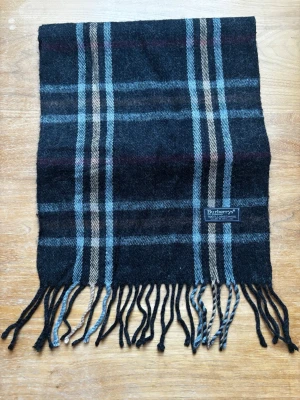 Burberrys halsduk - Snygg halsduk från Burberrys i svart, grått och blått rutmönster med inslag av vinrött. Gjord i mjuk ull och har klassiska fransar i ändarna. Perfekt accessoar för att lyfta din outfit.