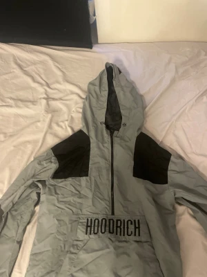 Grå vindjacka från Hoodrich - Snygg grå vindjacka från Hoodrich med svarta detaljer på axlar och huva. Jackan har dragkedja framtill, stor ficka med HOODRICH-tryck och luva med tryck baktill. Perfekt för streetwear och chill dagar.