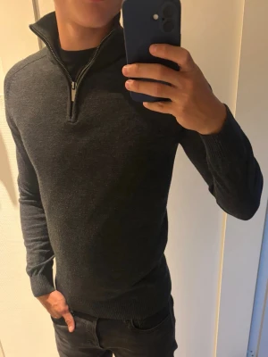 Mörkgrå stickad tröja med halvzip - Säljer min mörkgrå stickad tröja med halvzip och hög krage. Tröjan har långa ärmar och ribbade muddar vid ärmslut och nederkant. Skriv gärna vid intresse!