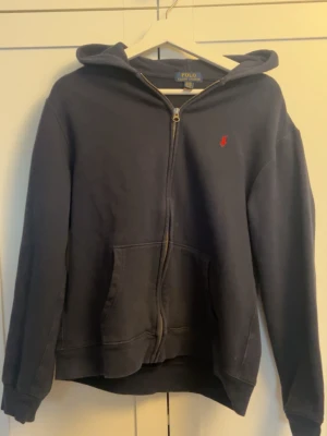 Mörkblå hoodie från Polo Ralph Lauren - Mörkblå hoodie med dragkedja från Polo Ralph Lauren. Klassisk design med huva, kängurufickor och den ikoniska röda logotypen broderad på bröstet. Tillverkad i mjukt bomullsmaterial som är skönt att bära. Perfekt för en avslappnad och stilren look.