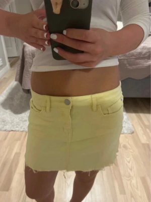 Gul jeanskjol med rå kant - Snygg kort gul jeanskjol med rå, fransig kant nertill och klassiska fickor både fram och bak. Kjolen har bälteshällor och stängs med knapp och dragkedja framtill. Perfekt för dig som vill ha en färgglad och trendig look.