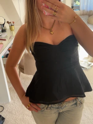 Svart axelbandslös peplum topp - Snygg svart axelbandslös topp med peplumskärning och lätt v-ringad byst. Toppen är figurnära upptill och har en utsvängd nederdel som ger en feminin siluett. Perfekt att styla med jeans eller kjol för en trendig look.