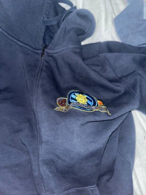 Mörkblå hoodie med broderad patch - Säljer en mörkblå hoodie med dragkedja och huva. På bröstet finns en broderad patch i blått, gult och rött med ett coolt emblem. Hoodien har dragsko i huvan och är gjord i mjukt sweatshirtmaterial. Perfekt för en avslappnad och stilren look.