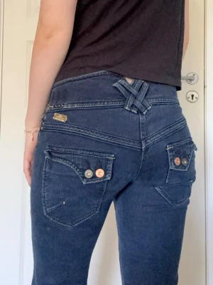 Low waist vintage jeans - Mörkblå low waist jeans från River Island med dubbla knappar fram, unika korsade detaljer bak i midjan och utsvängd passform. Midjemått rakt över 39cm och innerbenslängd 81cm💗