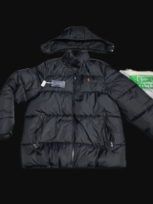 Svart dunjacka från Polo Ralph Lauren - Snygg svart dunjacka från Polo Ralph Lauren med avtagbar huva och dragkedja framtill. Jackan har två framfickor med dragkedja och den klassiska röda loggan på bröstet. Perfekt för kalla vinterdagar och riktigt stilren look.