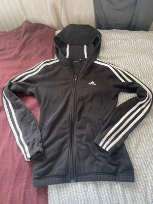 Svart Adidas hoodie med dragkedja - Snygg svart hoodie från Adidas med dragkedja framtill och klassiska vita ränder längs ärmarna. Tröjan har huva med vita detaljer och två fickor på sidorna. Perfekt för dig som gillar sportig och avslappnad stil.