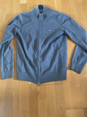 Grå ziptröja med hög krage - Snygg grå ziptröja med hög krage och ribbade muddar. Tröjan har hel dragkedja framtill och diskret broderad logga på bröstet. 