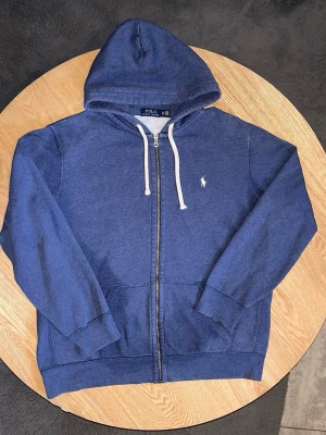 Polo Ralph Lauren Zip Hoodie Marinblå – Storlek XL - Säljer en Polo Ralph Lauren zip-hoodie i marinblå färg.  Klassisk modell med vit Polo-logo på bröstet, huva med vita snören och dragkedja framtill. Stilren och lättmatchad modell.  Storlek: XL (TG) Skick: Bra skick. Tvättad och klar.  Unisex: Ja Mått: Kan tas vid förfrågan.