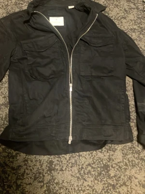 Svart overshirt jacka med dragkedja och fickor från Jack and Jones - Säljer en svart overshirt med rak passform och silverfärgad dragkedja framtill. Jackan har två stora bröstfickor med lock och lång ärm. Materialet ser ut att vara bomull eller en bomullsblandning. Perfekt för dig som vill ha en enkel och stilren overshirt. Är i storlek S men passar även M nästan aldrig använd de är bara att fråga om ni har funderingar! Nypris 799