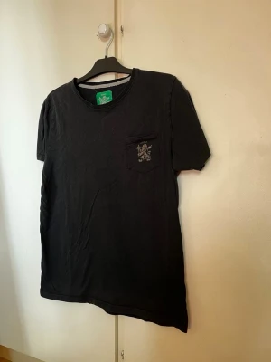 Åbro T-shirt  - Säljer en svart t-shirt med korta ärmar och rund hals. På bröstet finns en liten ficka med ett broderat Åbro lejonmärke. Perfekt till jeans eller shorts för en avslappnad look.