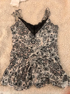 Söt volang topp - Snygg topp med tunna axelband, v-ringning och svart spetskant. Tyget är ljusrosa med svart blommigt och grafiskt mönster. Materialet är mjukt och stretchigt, vilket ger en bekväm passform och trendig look.