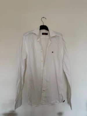 Vit långärmad Morris-skjorta - Klassisk vit skjorta med lång ärm och knappar framtill. Skjortan har en liten broderad logga på bröstet och en stilren krage. Perfekt för en clean och enkel look.