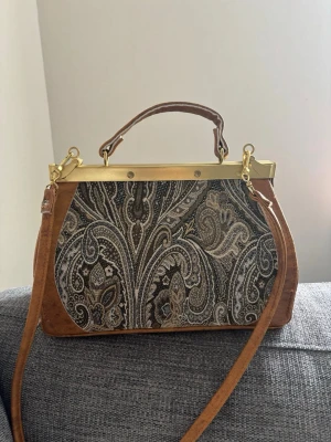 Brun paisleymönstrad handväska - Unik handväska med paisleymönstrad textil på framsidan och bruna läderdetaljer. Väskan har guldfärgade metalldetaljer, ett kort handtag och en avtagbar axelrem. Klassisk ramöppning ger en vintagekänsla och väskan har en mjuk, rundad form. Mått: 26x18x9 cm 