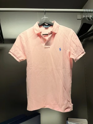 Ralph Lauren - Säljer en ljusrosa pikétröja från Ralph Lauren med klassisk krage och korta ärmar. Tröjan har den ikoniska blå loggan broderad på bröstet och är tillverkad i mjuk bomull. Perfekt för en avslappnad men ändå stilren look.