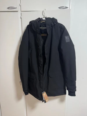 Svart parkas från Five Seasons - Snygg svart parkas från Five Seasons med huva och dragkedja framtill. Jackan har två stora fickor med lock och diskret logotyp på ärmen. Perfekt för kalla dagar och har en stilren, enkel design. Materialet är slitstarkt och vindtätt.