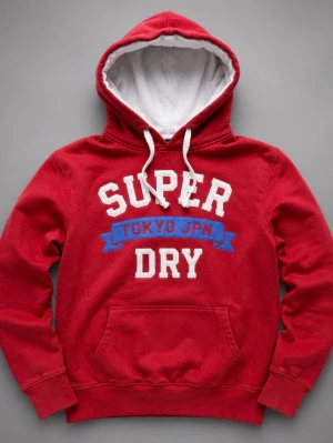 Röd Superdry hoodie med tryck - Röd hoodie från Superdry med vit och blå text på bröstet där det står 'SUPER DRY TOKYO JPN.'. Hoodien har vit dragsko, vit insida på huvan, dubbel huva och en stor magficka. Perfekt för en avslappnad streetstyle-look. Den är i storlek M, Hör av dig vid minsta lilla fundering!
