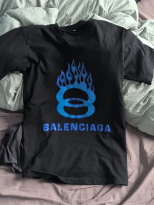 Svart t-shirt med blått tryck - Säljer en svart t-shirt med blått flames-tryck och logga på framsidan samt blå text på bak sidan.   Materialet känns som mjuk bomull och passformen är normal.