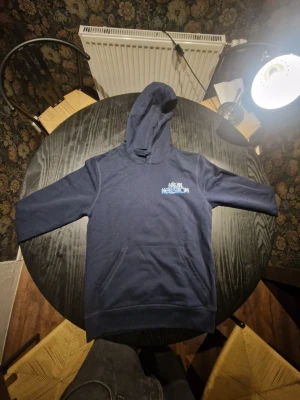 Mörkblå hoodie med tryck - Säljer en mörkblå hoodie med huva och känguruficka. På bröstet finns ett litet tryck med texten 'Håkan Hellström' i ljusblått och på ryggen ett stort färgglatt motiv av en person som sjunger, omgiven av stjärnor och sol. Perfekt för dig som gillar snygga och unika plagg.