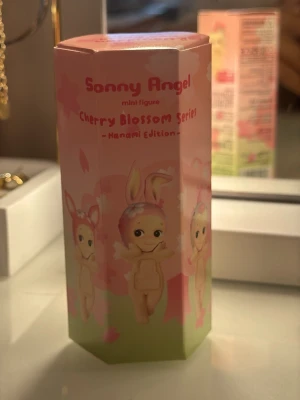 Sonny Angel Cherry Blossom Series - Säljer en Sonny Angel mini figure från Cherry Blossom Series Hanami Edition. Förpackningen är rosa och grön med söta illustrationer av figurer med olika djuröron och blomdetaljer. Perfekt för samlare eller som present. För mobilen!